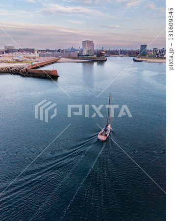 Aerial view of coastal harbor with sailboat leaving wake, flanked by breakwaters and dense city skyline blending maritime movement with urban infrastructure under muted skies. Aerial view of coastal harbor with sailboat leaving wake, flanked by breakwaters and dense city skyline blending maritime movement with urban infrastructure under muted skies. 131609345