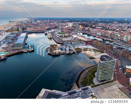 Aerial view of harbor city with curved docks, moored boats, and modern waterfront building framed by dense urban layout and sandy coastline under serene, partly cloudy skies. Aerial view of harbor city with curved docks, moored boats, and modern waterfront building framed by dense urban layout and sandy coastline under serene, partly cloudy skies. 131609350