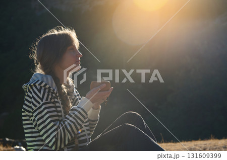 Golden Hour Outdoor Portrait Of A Woman In A Striped Hoodie Sitting On A Hill, Sipping From A Cup, Lost In Thought Amid Warm Sunset Light 131609399