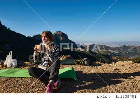 Woman Hiker Enjoying Snack On Mountain Summit With Backpack, Mat And Scenic Valley View 131609401