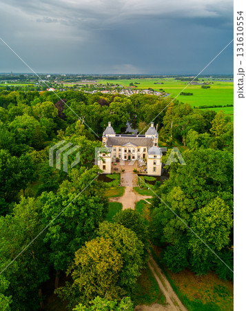 Aerial view of historic manor with twin towers, nestled in dense forest and open fields linked by a clear path and framed by distant village under dramatic overcast skies. 131610554