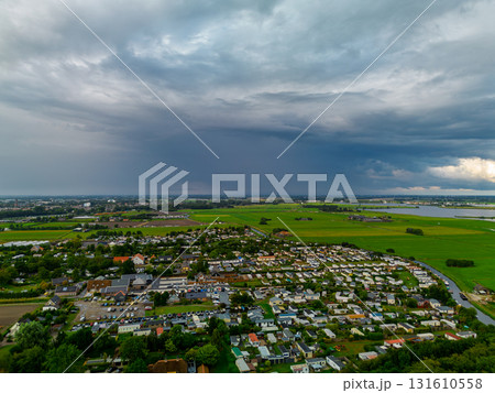 Aerial view of residential zone bordered by farmland and water dense housing, parked cars, and dramatic storm clouds contrast with vibrant green fields and rural openness. 131610558