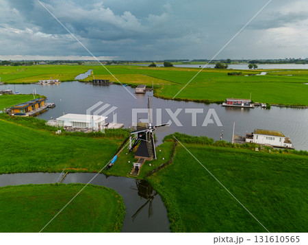 Aerial view of Dutch countryside with central windmill, houseboats, canal, and green fields capturing iconic rural charm and waterway culture under dramatic overcast skies. Aerial view of Dutch countryside with central windmill, houseboats, canal, and green fields capturing iconic rural charm and waterway culture under dramatic overcast skies. 131610565