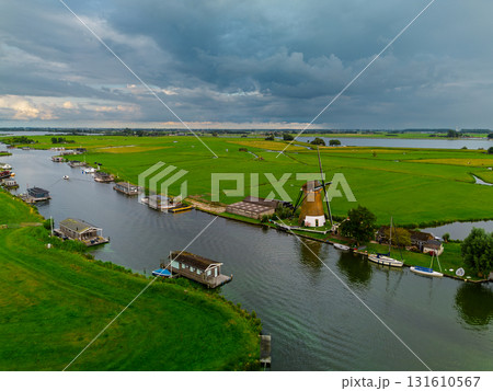Aerial view of Dutch countryside with traditional windmill, canal, docked boats, and green fields capturing rural charm and water management under dramatic overcast skies. Aerial view of Dutch countryside with traditional windmill, canal, docked boats, and green fields capturing rural charm and water management under dramatic overcast skies. 131610567