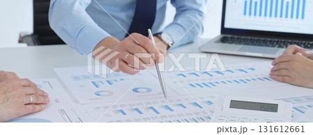 Business professional pointing at printed charts with a pen during a meeting, laptop showing bar graphs and calculator on table while team collaborates on financial analysis and planning 131612661