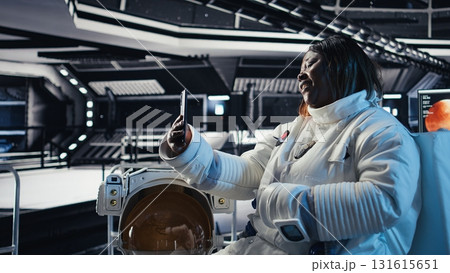Happy female cosmonaut doing videocall with family using smartphone during galaxy expedition. Africna american woman doing remote call with loved ones using mobile phone in spaceship Happy female cosmonaut doing videocall with family using smartphone during galaxy expedition. Africna american woman doing remote call with loved ones using mobile phone in spaceship 131615651