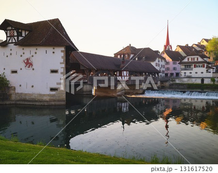 Historic wooden bridge and charming buildings reflect in calm water during golden hour in picturesque Swiss town 131617520