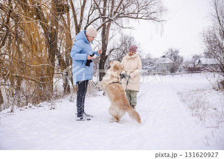 Elderly woman and her adult daughter walking with a Golden Retriever on a snowy rural path. The dog stands on its hind legs near the younger woman while snow falls around them in a peaceful winter Elderly woman and her adult daughter walking with a Golden Retriever on a snowy rural path. The dog stands on its hind legs near the younger woman while snow falls around them in a peaceful winter 131620927