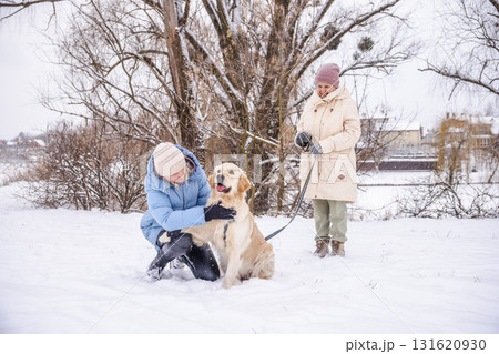 Elderly woman and her adult daughter enjoying a winter walk with their Golden Retriever. The daughter kneels to pet the happy dog while the mother holds the leash, surrounded by snowy trees Elderly woman and her adult daughter enjoying a winter walk with their Golden Retriever. The daughter kneels to pet the happy dog while the mother holds the leash, surrounded by snowy trees 131620930