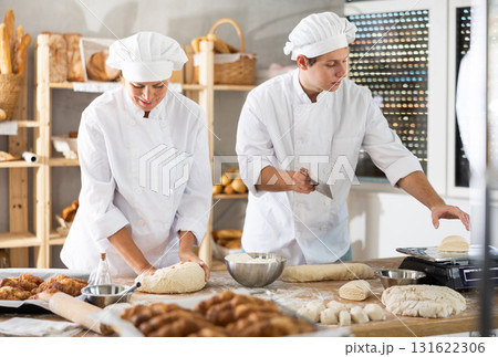 Female baker kneads dough while young guy cuts pieces of raw dough and weighs them on scales in bakery kitchen Female baker kneads dough while young guy cuts pieces of raw dough and weighs them on scales in bakery kitchen 131622306