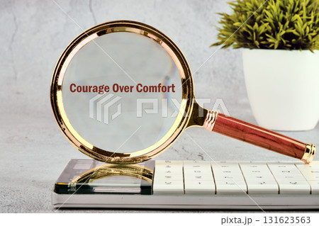 A magnifying glass rests on a calculator. Through the lens, you can see the words Courage Over Comfort. A potted plant adds a touch of nature to the workspace A magnifying glass rests on a calculator. Through the lens, you can see the words Courage Over Comfort. A potted plant adds a touch of nature to the workspace 131623563