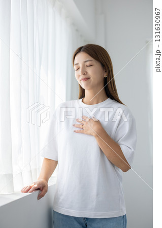 明るい窓辺で胸に手を当てる若い女性 明るい窓辺で胸に手を当てる若い女性 131630967