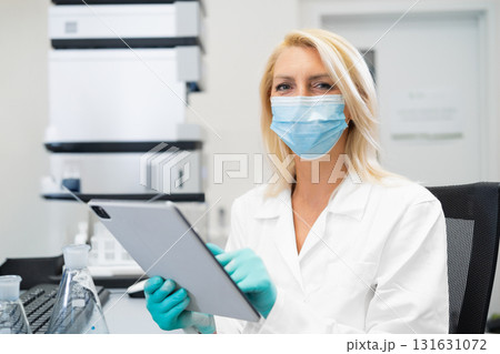 Blonde female scientist wearing a lab mask and rubber gloves recording chemical experiment results on a tablet while standing in a chemistry laboratory with instruments around. Blonde female scientist wearing a lab mask and rubber gloves recording chemical experiment results on a tablet while standing in a chemistry laboratory with instruments around. 131631072