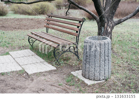An empty bench with wooden seating planks and a trash can under a tree in a city park. A modern wrought iron bench and trash can sit on the edge of a city park path on a spring day. An empty bench with wooden seating planks and a trash can under a tree in a city park. A modern wrought iron bench and trash can sit on the edge of a city park path on a spring day. 131633155
