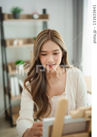 Young woman with long hair engaged in painting on canvas in a cozy, bright living space with wooden shelves and green plants in the background Young woman with long hair engaged in painting on canvas in a cozy, bright living space with wooden shelves and green plants in the background 131633641