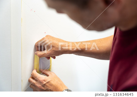 Man covering a light switch with masking tape before painting the wall white. Home renovation, surface protection and careful preparation during interior work. Man covering a light switch with masking tape before painting the wall white. Home renovation, surface protection and careful preparation during interior work. 131634645