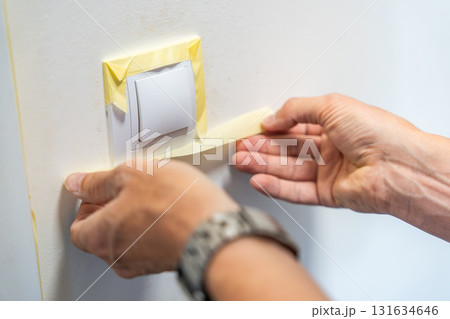 Man covering a light switch with masking tape before painting the wall white. Home renovation, surface protection and careful preparation during interior work. Man covering a light switch with masking tape before painting the wall white. Home renovation, surface protection and careful preparation during interior work. 131634646