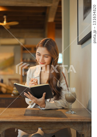 Smiling businesswoman in beige blazer sitting at a wooden table, taking notes in a notebook 131634820