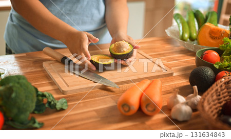 Hands cutting ripe avocado on wooden cutting board surrounded by fresh vegetables Hands cutting ripe avocado on wooden cutting board surrounded by fresh vegetables 131634843