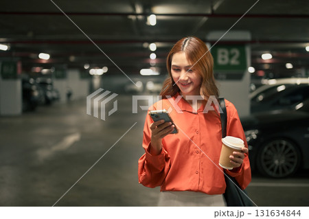 Smiling woman standing in parking garage using smartphone while holding coffee cup 131634844
