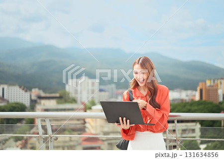 Smiling young woman using laptop outdoors on rooftop with city and mountain view Smiling young woman using laptop outdoors on rooftop with city and mountain view 131634856