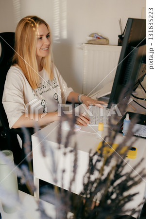 Woman working from home, sitting comfortably at bright white desk, typing on sleek white keyboard while smiling and engaging with computer screen in her cozy workspace. Professional blonde freelancer 131635223