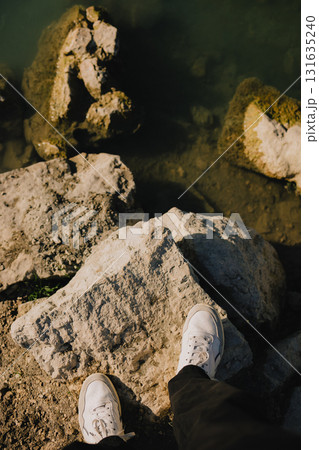First-person view: person standing on large rocks by tranquil body of water, wearing white sneakers and black pants, soaking in the vibrant outdoor scenery on bright, sunny day. Men legs in sneakers 131635240