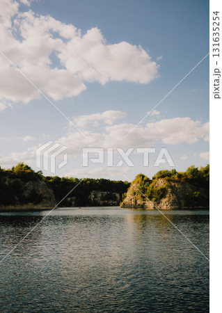 Calm lake water reflecting bright sky filled with fluffy white clouds, surrounded by rocky hills adorned with lush vegetation on sunny summer day, creating serene landscape. Tranquil lake. Nature Calm lake water reflecting bright sky filled with fluffy white clouds, surrounded by rocky hills adorned with lush vegetation on sunny summer day, creating serene landscape. Tranquil lake. Nature 131635254