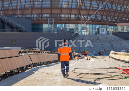 Worker in safety gear walking through construction site near modern building. construction worker in orange and blue uniform walks away from camera at building site with construction materials Worker in safety gear walking through construction site near modern building. construction worker in orange and blue uniform walks away from camera at building site with construction materials 131635362
