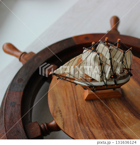 A scale model of an old sailing ship sits on the wooden center of a ship's steering wheel on a shelf  131636124