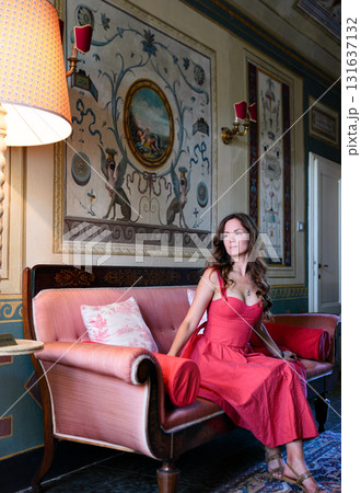 Woman in coral dress sitting on pink sofa in classic interior Woman in coral dress sitting on pink sofa in classic interior 131637132