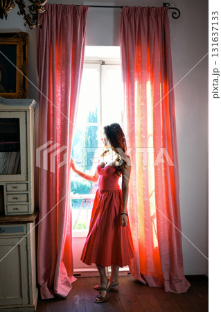 Woman in coral dress standing by window with pink curtains Woman in coral dress standing by window with pink curtains 131637133