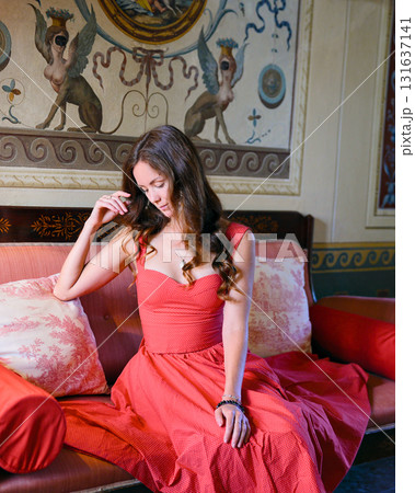 Woman in coral dress sitting on bed in classic interior Woman in coral dress sitting on bed in classic interior 131637141