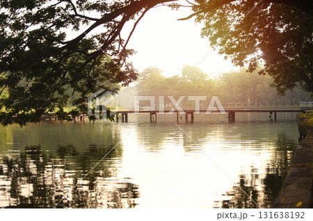 A wooden bridge across a pond in the park on a misty morning. A quiet place to relax. Still warm orange from the sun, there are large branches in the foreground. A wooden bridge across a pond in the park on a misty morning. A quiet place to relax. Still warm orange from the sun, there are large branches in the foreground. 131638192