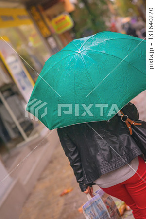 Woman rushing with green teal umbrella on rainy urban street. Intentional rotation of angle towards horizon. Concept of autumn weather, lifestyle, city rain mood, street life in motion 131640220