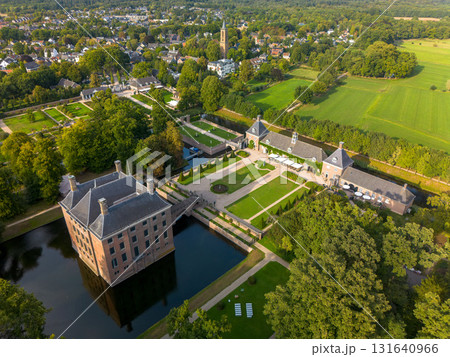 Aerial view of historic moated castle with gardens, bridge, and adjacent long building, surrounded by trees, water features, and a nearby village under warm sunlight. 131640966