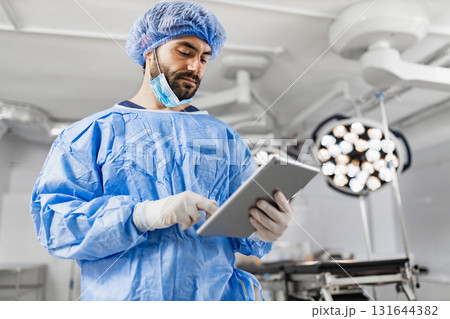 A surgeon in scrubs and a surgical cap uses a tablet in a modern operating room, likely reviewing patient information or surgical plans. A surgeon in scrubs and a surgical cap uses a tablet in a modern operating room, likely reviewing patient information or surgical plans. 131644382