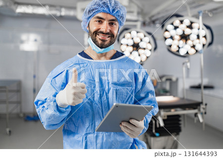 A smiling surgeon in scrubs gives a thumbs up while holding a tablet in an operating room. 131644393