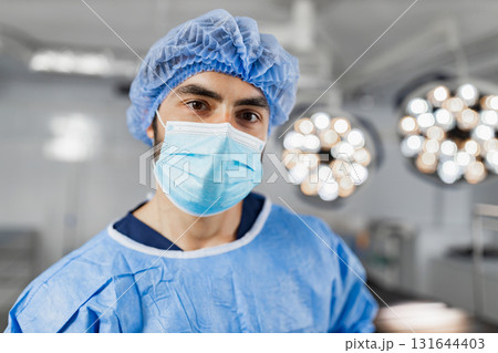 A surgeon wearing a surgical mask and cap stands in an operating room, illuminated by bright lights. 131644403