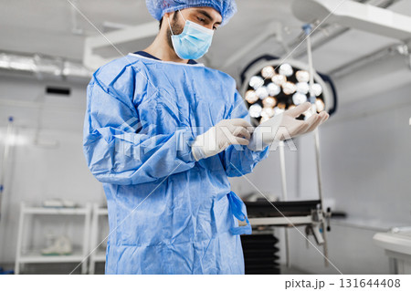 A surgeon in an operating room puts on sterile gloves, preparing for a medical procedure. The scene is well-lit, highlighting the surgical environment. 131644408