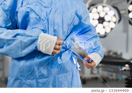 A surgeon prepares for an operation, tying the back of their blue surgical gown in a sterile operating room setting. 131644409