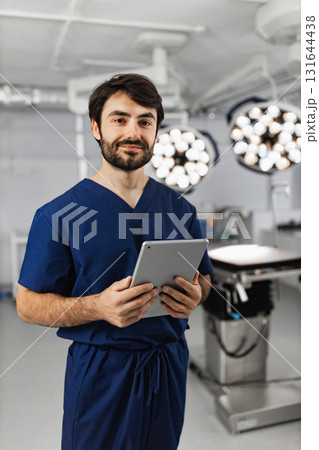 A surgeon stands in an operating room, holding a tablet and smiling at the camera. 131644438