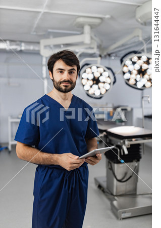 A surgeon in blue scrubs stands in an operating room, holding a tablet and looking at the camera. A surgeon in blue scrubs stands in an operating room, holding a tablet and looking at the camera. 131644447
