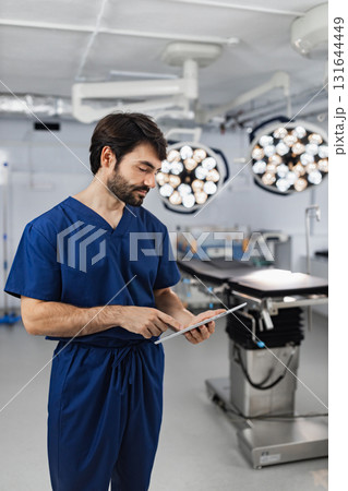 A surgeon in blue scrubs reviews a tablet in a modern operating room, with surgical lights and equipment in the background. 131644449