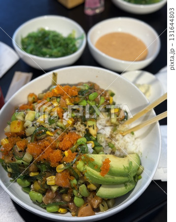 Bowl filled with sushi rice, topped with avocado, edamame, corn, and fish roe, surrounded by dipping sauces and green onions, highlighting delicious food presentation 131644803