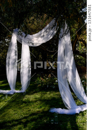 Outdoor wedding ceremony with a white fabric arch among trees and on grass Outdoor wedding ceremony with a white fabric arch among trees and on grass 131647881