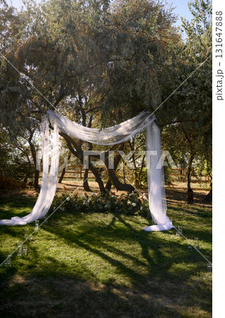 Outdoor wedding ceremony with a white fabric arch among trees and on grass Outdoor wedding ceremony with a white fabric arch among trees and on grass 131647888