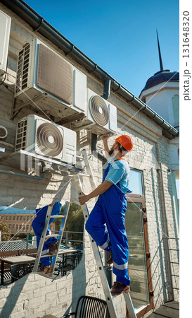 Worker on ladder maintaining multiple air conditioning units on building wall Worker on ladder maintaining multiple air conditioning units on building wall 131648320