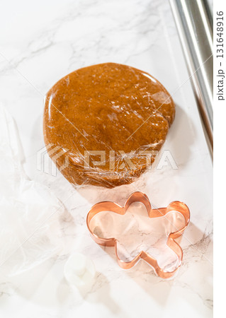 Diagonal view of wrapped gingerbread dough next to a rolling pin and gingerbread man cookie cutters on a marble surface. 131648916