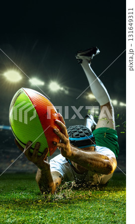 Rugby player diving across 3D grass field holding colorful ball under bright lights 131649311